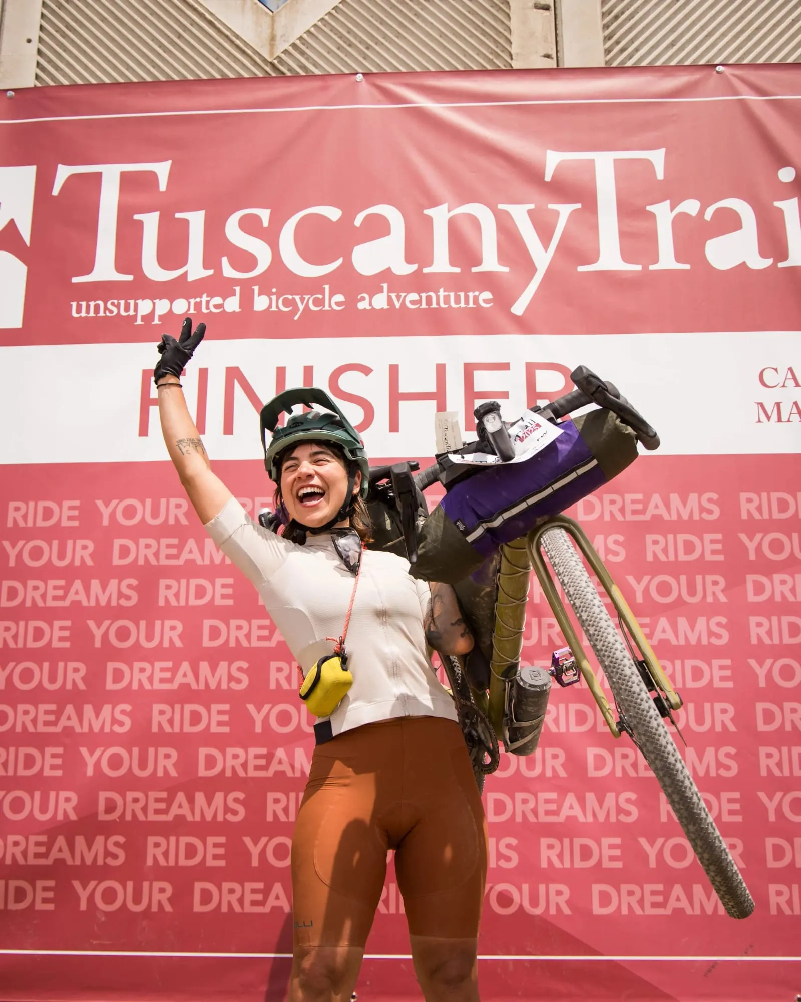 Una finisher alza la bici sopra la testa davanti allo striscione Ride your dreams