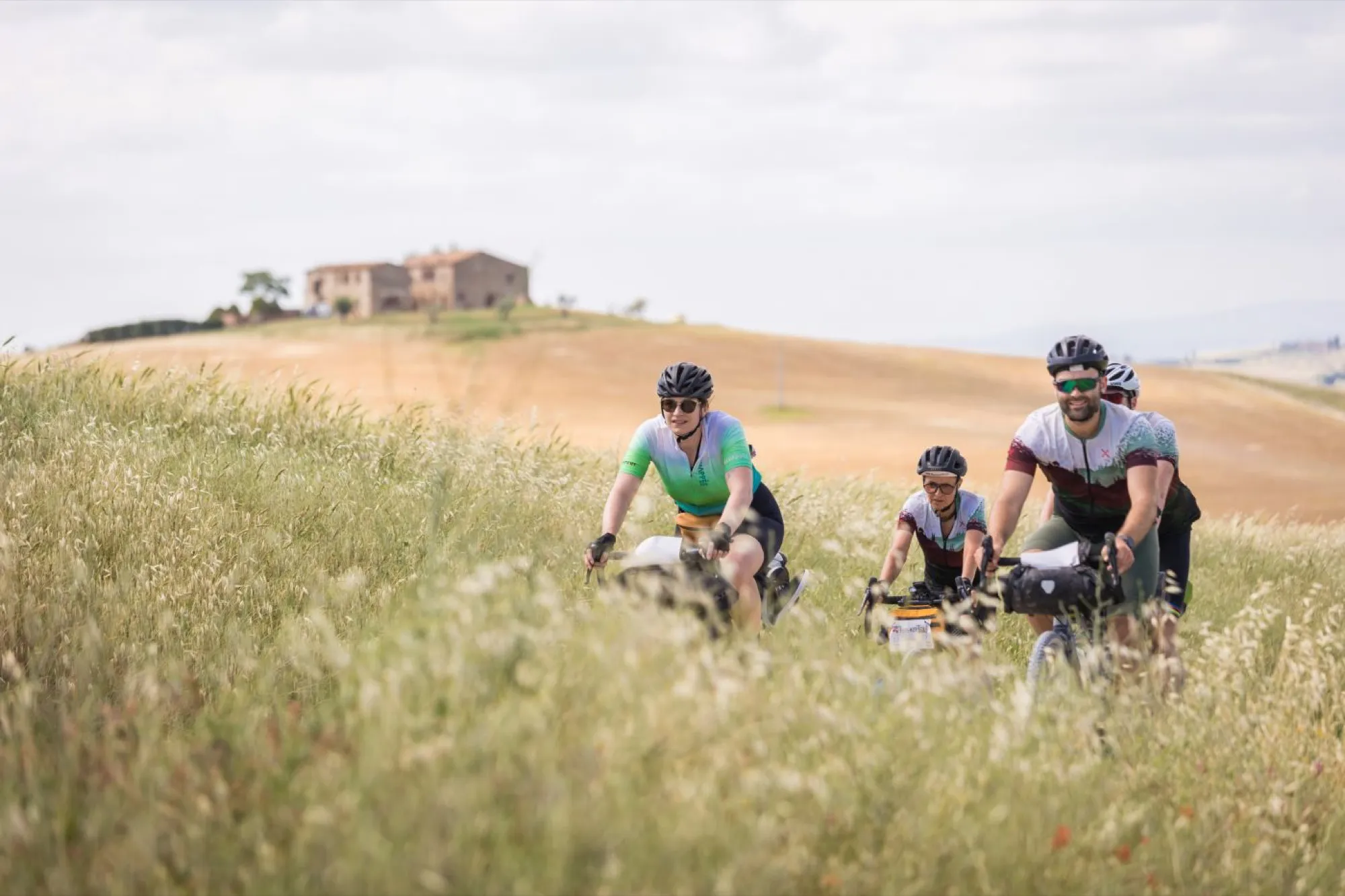 Tre rider con le borse attraversano un campo di spighe, un casale sulla collina